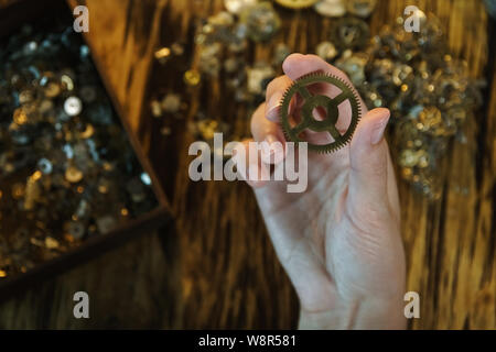 Orologiaio detiene il grande ingranaggio. Ruota dentata su sfondo sfocato di workplace. Pignone in mano femminile. Vecchia ruota dentata sul tavolo di legno dello sfondo. Metallo grande ge Foto Stock