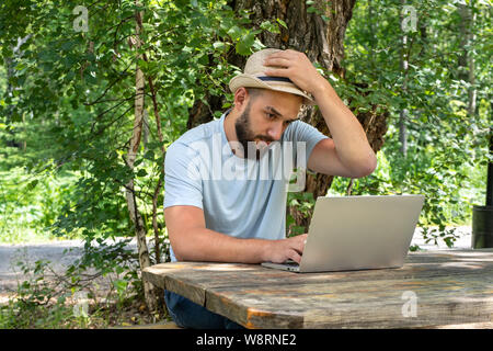 Uomo Barbuto è perplesso e pensa che mentre si lavora su un computer portatile all'aperto presso una tabella nel parco. Il concetto di lavoro remoto, office, freelance Foto Stock