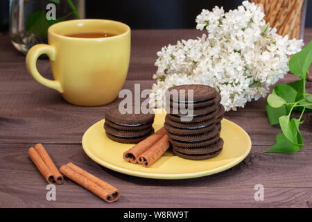 Coppia di tè e coppa giallo piattino. I biscotti al cioccolato, bastoncini di cannella, lilla bianco al buio su un tavolo di legno della cerimonia del tè, piccoli snack, dessert Foto Stock