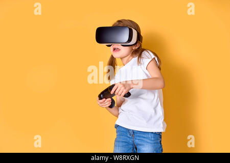 Ragazza sperimentando auricolare VR vs joystick gioco su sfondo giallo. Sorpreso emozioni sul suo viso. Bambino usando un gadget di gioco di realtà virtuale. Occhiali futuristici in giovane età. La tecnologia virtuale Foto Stock