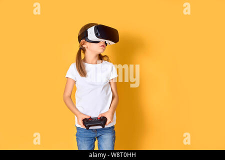 Ragazza sperimentando auricolare VR vs joystick gioco su sfondo giallo. Sorpreso emozioni sul suo viso. Bambino usando un gadget di gioco di realtà virtuale. Occhiali futuristici in giovane età. La tecnologia virtuale Foto Stock