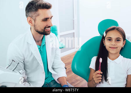 Po' di razza mista ragazza in poltrona odontoiatrica seduto vicino al suo dentista. Ella holding spazzolino e sorridente. Il modo giusto per spazzolare i denti Foto Stock