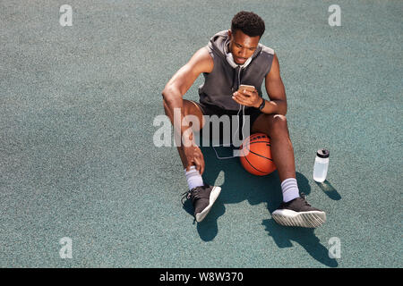 Al di sopra di vista sullo sfondo di un bel afro-americano di uomo seduto in un campo da pallacanestro e utilizza lo smartphone, spazio di copia Foto Stock