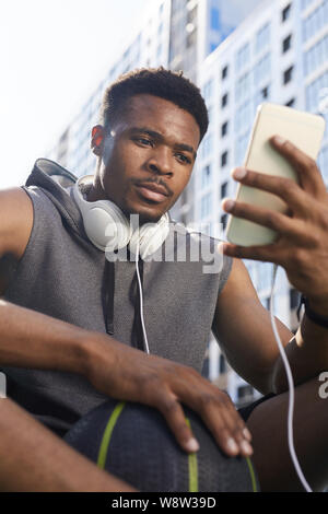 Angolo basso ritratto di contemporaneo afro-uomo utilizza lo smartphone mentre è seduto nella pallacanestro, spazio di copia Foto Stock