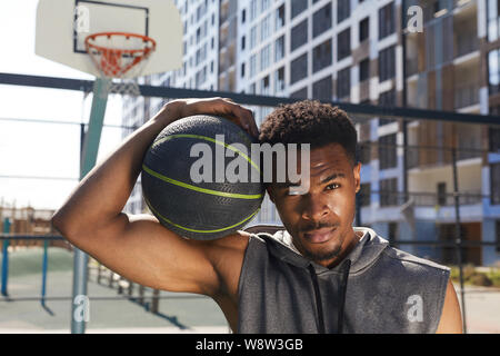 Ritratto di bello afro-americano di uomo con il basket ball guardando la fotocamera mentre in posa nella corte di sport all'aperto, spazio di copia