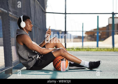 Vista laterale verticale del contemporaneo afro-uomo che indossa le cuffie e utilizza lo smartphone mentre sedendo nel campo da basket all'aperto, spazio di copia Foto Stock