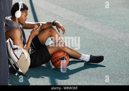 Angolo alto ritratto di contemporaneo afro-uomo che indossa le cuffie e utilizza lo smartphone mentre sedendo nel campo da basket all'aperto, spazio di copia Foto Stock