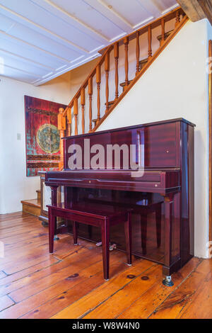 Pianoforte bordeaux verticale accanto alla scala in legno di pino che conduce al piano superiore all'interno di una vecchia casa in pietra fieldstone stile cottage del 1820. Foto Stock