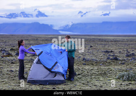 Tenda - persone pitching tenda in Islanda al crepuscolo. Paio di impostazione di campo di notte dopo le escursioni nella selvaggia natura Islandese paesaggio. Asia multiculturale della donna e uomo caucasico uno stile di vita sano. Foto Stock