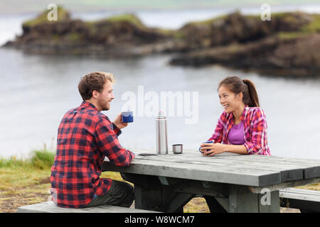 Camping giovane seduta a tavola di bere il caffè dal thermos pallone dal lago in Islanda. Camper donna e uomo prendendo rilassante pausa sul viaggio nella splendida natura Islandese. Foto Stock