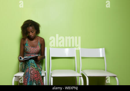 Bella americano africano donna seduta su una sedia e la lettura rivista nella sala di attesa dell'ospedale Foto Stock