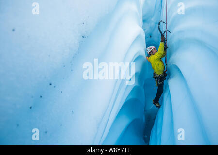 Uomo in cappotto giallo arrampicata su ghiaccio una stretta e canyon verticale tagliato dal solido ghiaccio del ghiacciaio. Foto Stock