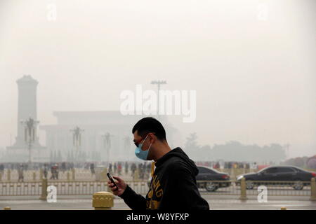 Un uomo che indossa una maschera facciale usa il suo smartphone come egli cammina oltre la piazza Tiananmen in pesanti smog a Pechino, in Cina, il 9 ottobre 2014. I piani di Pechino t Foto Stock