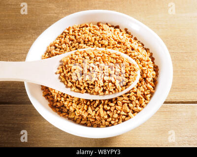 Fenugreek (Trigonella) in tazza bianca su un cucchiaio di legno su un sfondo di legno Foto Stock