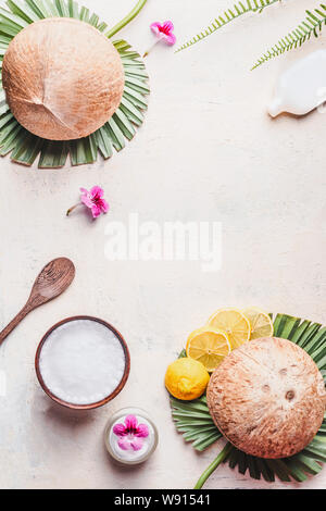 Olio di noce di cocco nella ciotola di legno con cucchiaio e crema idratante sulla scrivania bianca di sfondo tropicale con foglie e fiori , vista dall'alto. Telaio. Copia dello spazio. Foto Stock