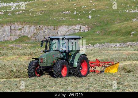 Agricoltore remare fino al fieno con un trattore Hurlimann e a due pass rastrello su una collina di pennini agriturismo vicino a Ravenstondeale, Cumbria. Foto Stock