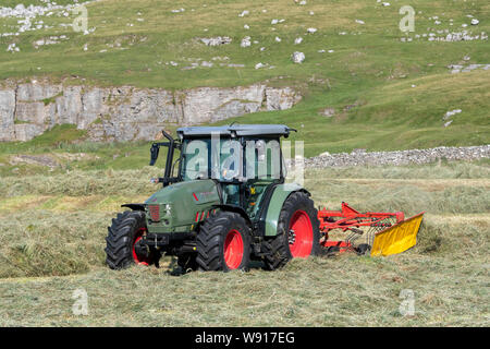 Agricoltore remare fino al fieno con un trattore Hurlimann e a due pass rastrello su una collina di pennini agriturismo vicino a Ravenstondeale, Cumbria. Foto Stock