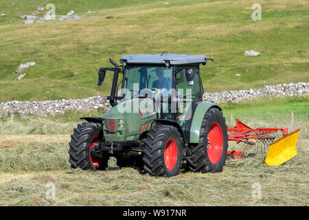 Agricoltore remare fino al fieno con un trattore Hurlimann e a due pass rastrello su una collina di pennini agriturismo vicino a Ravenstondeale, Cumbria. Foto Stock