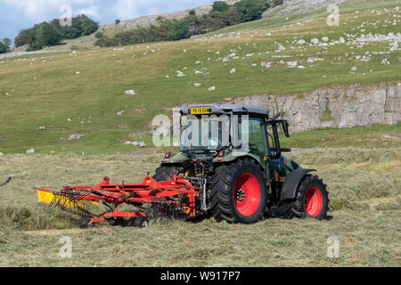 Agricoltore remare fino al fieno con un trattore Hurlimann e a due pass rastrello su una collina di pennini agriturismo vicino a Ravenstondeale, Cumbria. Foto Stock