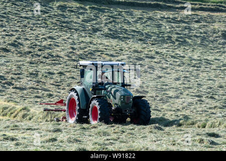 Agricoltore remare fino al fieno con un trattore Hurlimann e a due pass rastrello su una collina di pennini agriturismo vicino a Ravenstondeale, Cumbria. Foto Stock