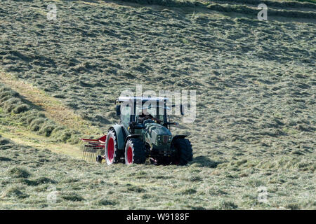 Agricoltore remare fino al fieno con un trattore Hurlimann e a due pass rastrello su una collina di pennini agriturismo vicino a Ravenstondeale, Cumbria. Foto Stock