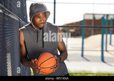 Vita ritratto di contemporaneo afro-uomo holding basket ball in posa nella corte di sport all'aperto, spazio di copia Foto Stock