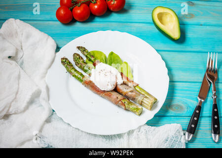 Asparagi verdi grigliati avvolto con pancetta, benedetto uovo in camicia blu su uno sfondo di legno. Prima colazione Foto Stock