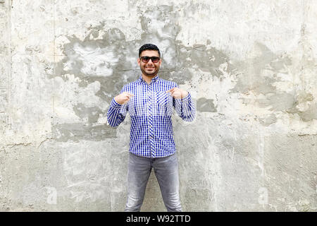 Questo è me. Ritratto di felice bello barbuto giovane a scacchi in maglietta blu, occhiali da sole in piedi contro il cemento grigio a parete, di puntamento e di mostra Foto Stock