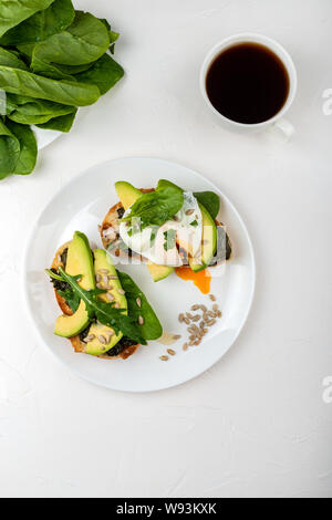 La prima colazione. Crostini di pane con uovo in camicia e avocado con spinaci freschi rucola e semi e una tazza di caffè Foto Stock