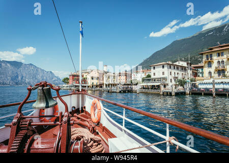 Prua di una barca con campana nave per una crociera tour. L'acqua è blu e la gamma della montagna e piccolo grazioso villaggio al Lago di Garda, Italia Foto Stock