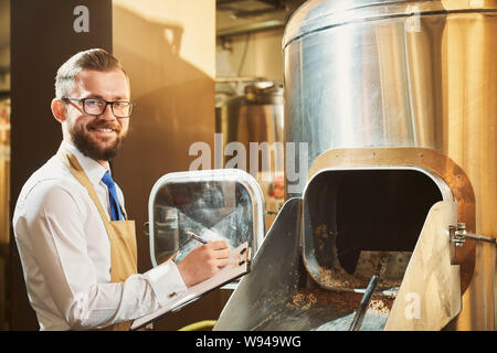 Allegro birraio maschio che indossano occhiali guardando la fotocamera e sorridere mentre la scrittura dei dati nella cartella. Uomo di esaminare la qualità di grist e malto presso la fabbrica di produzione. Concetto di birreria. Foto Stock