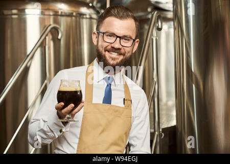 Vista frontale di felice barbuto birraio maschio mantenendo il bicchiere di birra scura in mano, guardando la fotocamera e sorridente. Uomo che lavora nella fabbrica di birra e di esaminare la qualità di ale. Concetto di produzione. Foto Stock