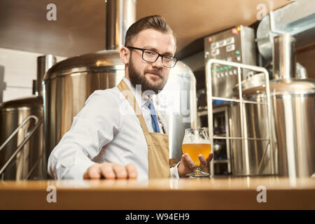 Giovani barbuto brewer guardando la fotocamera e sorridente mantenendo un bicchiere di ale dorata in mano. Uomo che indossa una camicia bianca e grembiule in piedi in birreria e esaminando la birra. Concetto di produzione. Foto Stock