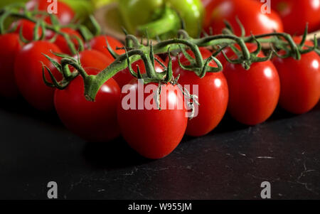 Closeup, piccoli pomodori ciliegia con foglie di vite, offuscata pepe verde in background, sul marmo scuro come il bordo Foto Stock