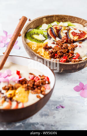 Ciotola frullato con frutta e muesli con guscio di noce di cocco ciotola su sfondo grigio. Sano cibo vegan concetto. Foto Stock