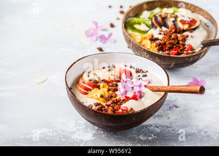 Ciotola frullato con frutta e muesli con guscio di noce di cocco ciotola su sfondo grigio. Sano cibo vegan concetto. Foto Stock