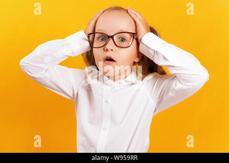Ritratto di un allegro bambina in bicchieri su sfondo giallo. Il bambino è sconvolto e tiene le mani dietro la testa. Foto Stock