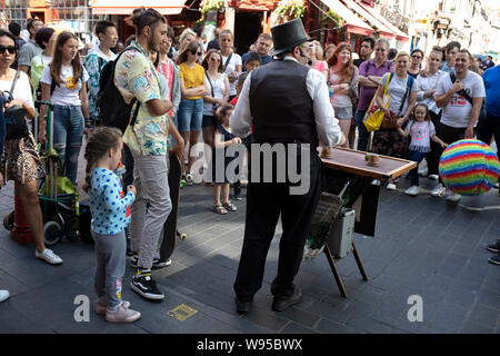 Scena di strada come una folla di turisti di tutte le età si riuniscono per guardare una strada magic show da un esecutore di strada / busker indossando un waitcoat e un cappello a cilindro nella Chinatown di Soho, Londra, Regno Unito. La presente Chinatown si trova nella zona di Soho che occupa l'area in e attorno a Gerrard Street. Esso contiene un certo numero di ristoranti cinesi, panifici, supermercati, negozi di souvenir e altri cinese-gestione delle imprese ed è di per sé un importante destinazione turistica. Foto Stock