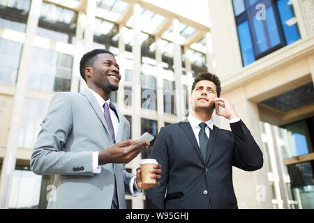 Due business di successo di manager in giacca utilizzando i telefoni cellulari e occupato la conversazione mentre passeggiate in città Foto Stock