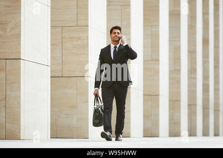 Giovane imprenditore in abito nero borsa da trasporto e ha una conversazione aziendale sul telefono cellulare mentre passeggiate in città Foto Stock