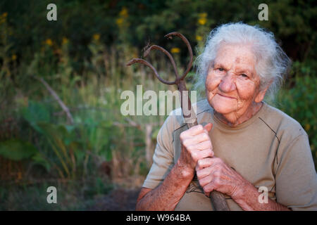 Closeup ritratto di una vecchia donna con i capelli grigi tenendo un arrugginito forcone o il tritatutto nelle sue mani, faccia in profonde rughe, il fuoco selettivo Foto Stock