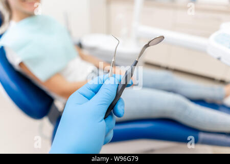 Close-up mano del dentista in guanto contiene tool sonda e specchio. Il paziente nel riunito dentale in background. Un lavoro dentario in clinica. Office Foto Stock