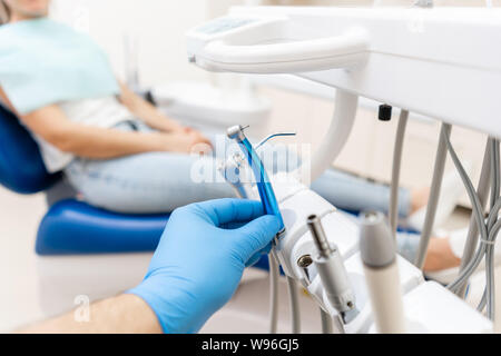 Close-up mano del dentista in guanto contiene dental turbina ad alta velocità. Il paziente in sedia blu sullo sfondo. Ufficio dove dentista conduce Foto Stock