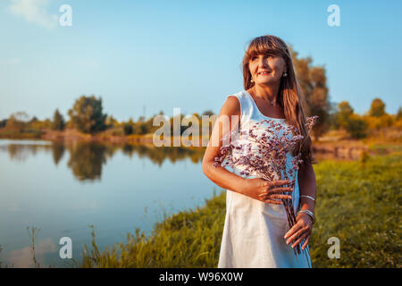 Donna matura azienda bouquet di fiori di fronte al fiume in autunno park. Senior donna ammira il paesaggio Foto Stock