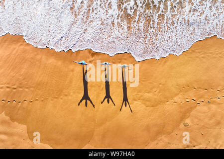 Tre persone che guardano verso il rotolamento onde dell oceano Pacifico su una superficie pulita ampia spiaggia di sabbia durante la ginnastica mattutina di antenna in vista dall'alto in basso. Foto Stock