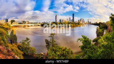 Verdi scogliere fresco parco sulle rive del fiume Brisbane attraverso città CBD alti torri in ampio panorama sulla mattina di sole sotto il cielo blu. Foto Stock