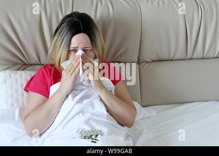 Donna soffia il naso in un fazzoletto di seduta in un letto. Infelice fanciulla soffre di naso che cola, concetto di malati a casa, raffreddori e influenza, febbre Foto Stock