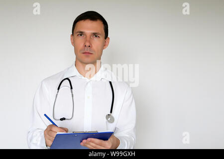 Medico con stetoscopio scrive una prescrizione in piedi su sfondo bianco. Ritratto di un medico di sesso maschile, il concetto di medicina, terapeuta, esame Foto Stock