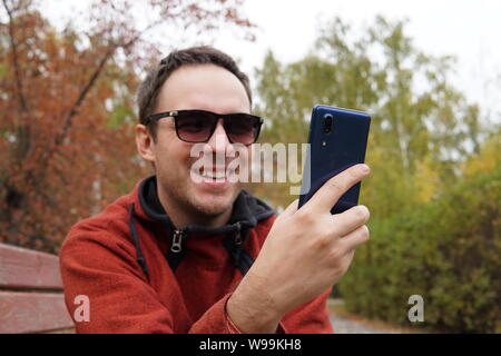 Giovane uomo seduto in posizione di parcheggio e utilizzo di smart phone. Sorridenti guy la lettura di un messaggio sul cellulare mentre è seduto su una panchina nel parco. l uomo surf net Foto Stock