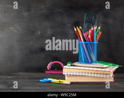 Pila di notebook, vetro blu con multi-di legno colorato Matite e penne, righelli e una gomma sullo sfondo di un vuoto di gesso nero bordo, indietro Foto Stock